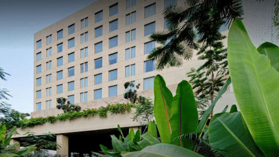 Exterior facade of Polo Hotel, Agartala showing the multi-story beige building with tinted blue windows, lush tropical foliage and banana plants framing entrance.