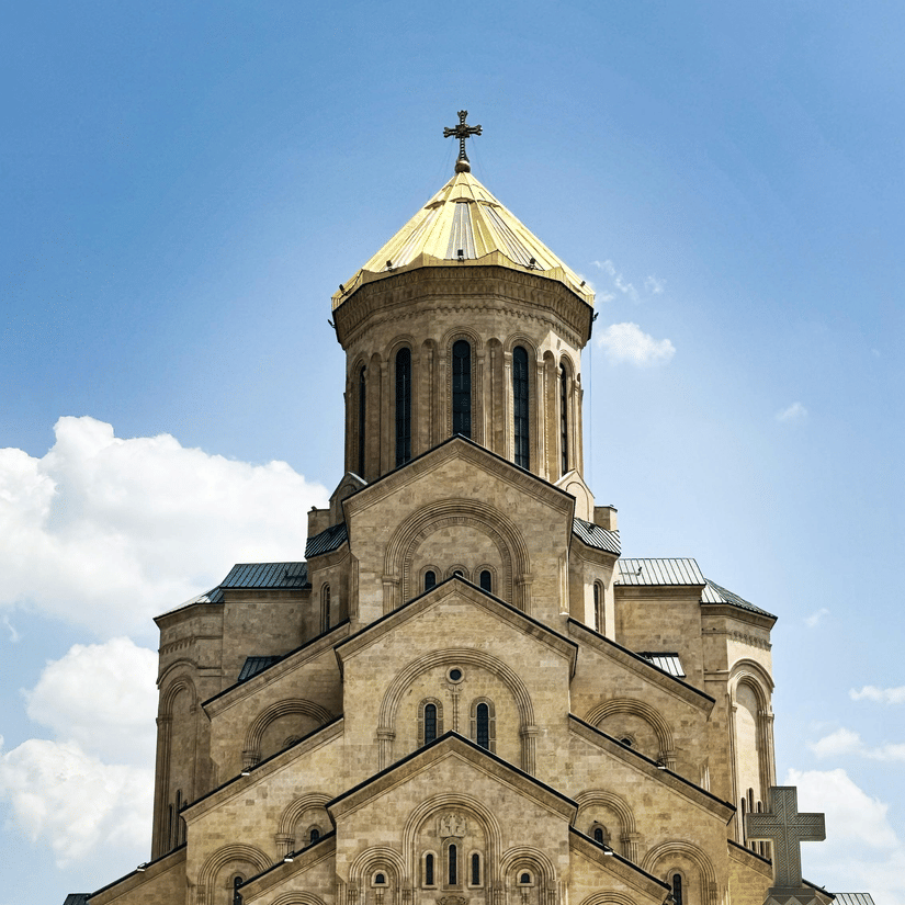 A large, multi-tiered stone cathedral featuring a golden dome and cross set against a bright, slightly cloudy blue sky.