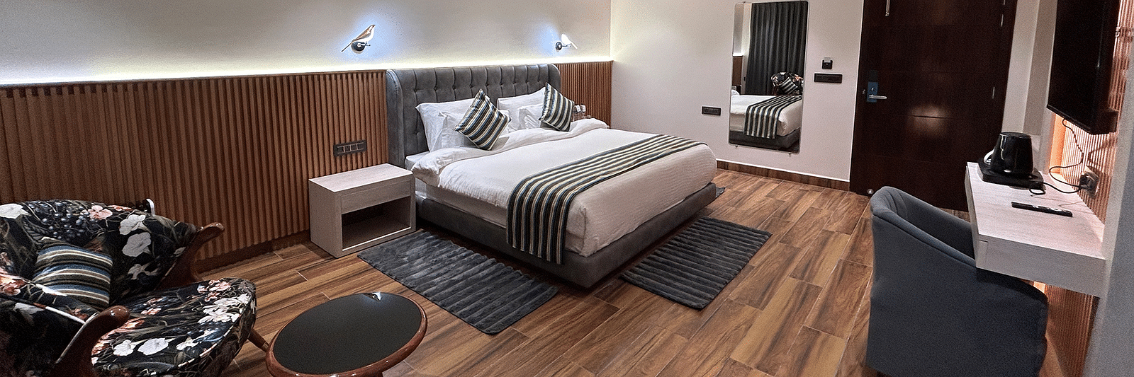 A shot of the deluxe room featuring a bed, bed side tables, two chairs, coffee table, work desk, a couch, a flat screen TV, and a full length mirror - DLS The Emerald, Nainital