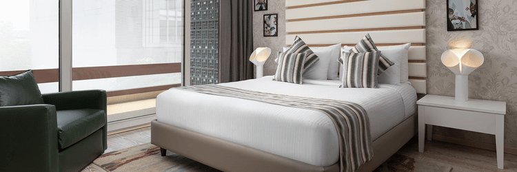 A stylish room at The Atara, Gurugram boasts a comfortable bed with a patterned runner, a plush green armchair, and a large window that offers a scenic outdoor view