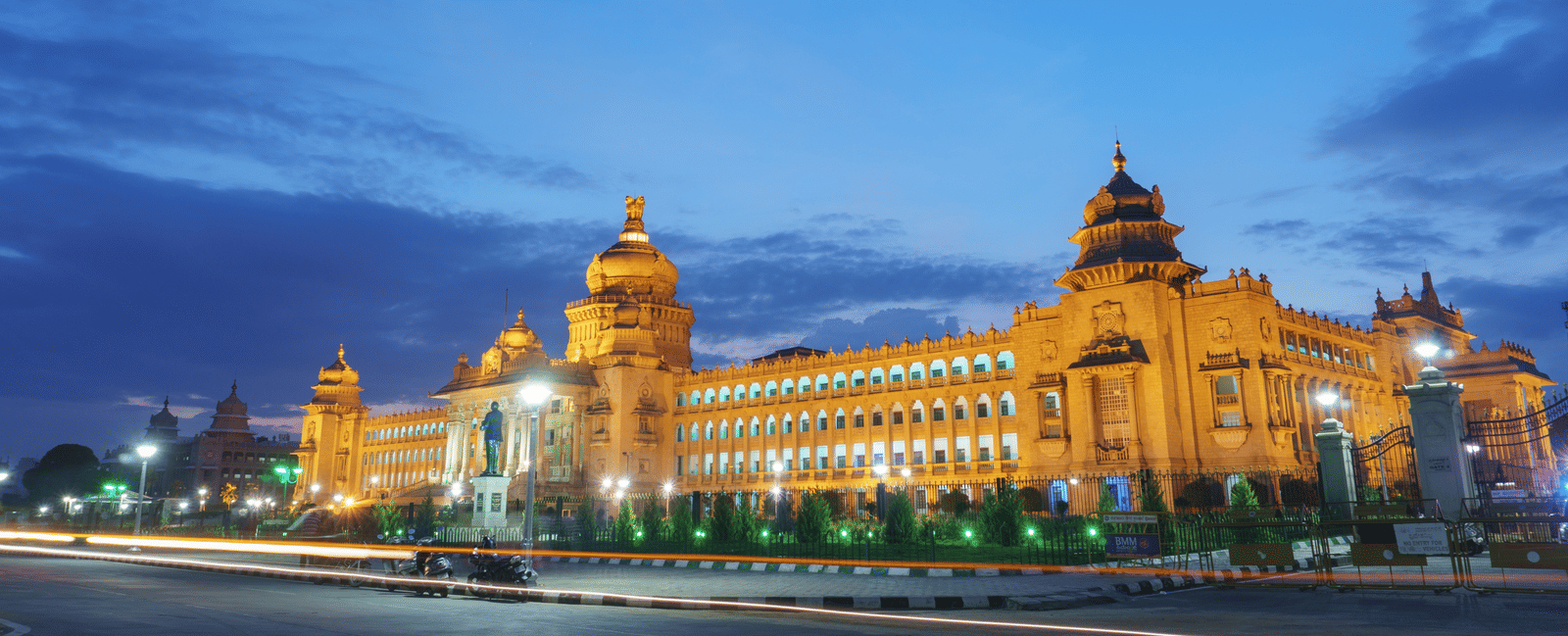 Facade of the Vidhana Soudha illuminated with warm lights at dusk and two bikes parked by the road in front
