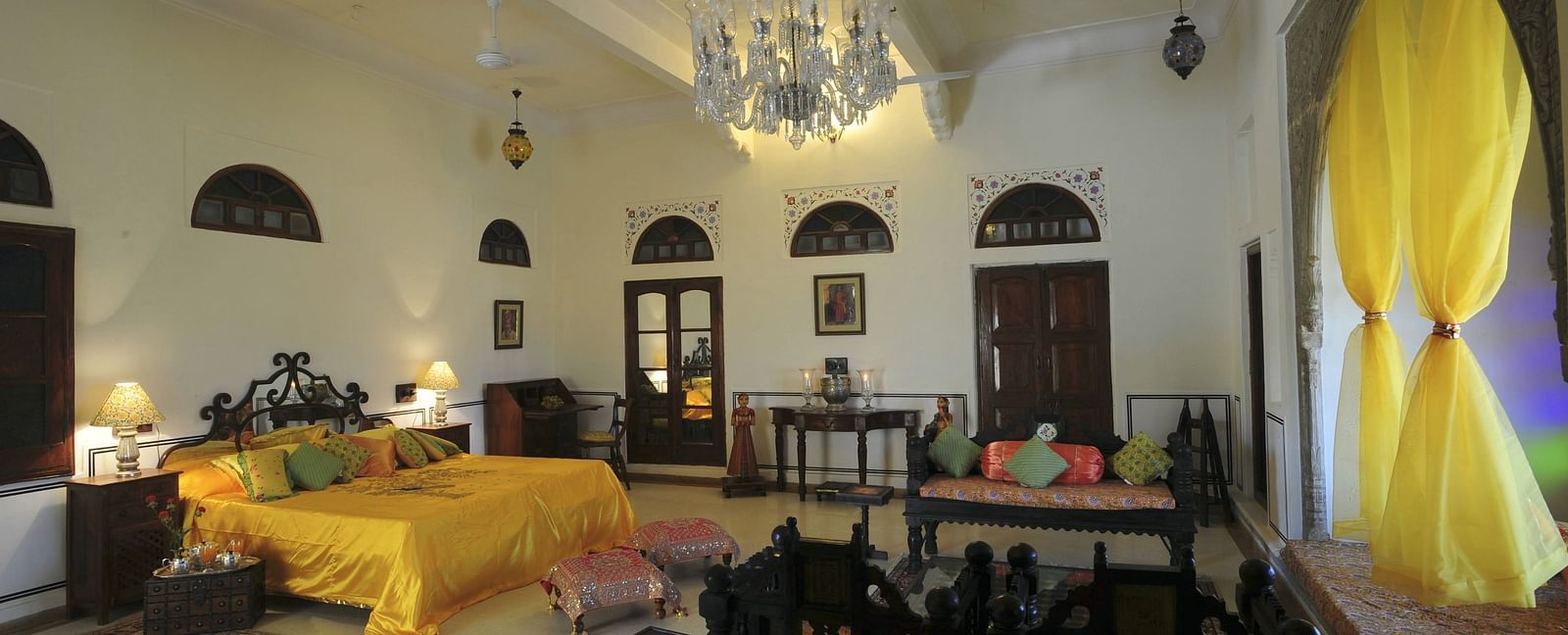 A spacious bedroom at Fort Barli, featuring a vibrant yellow bedspread, a crystal chandelier, and antique furniture.
