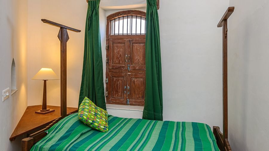Room with bed, striped bedding, patterned pillow, lamp on table, curtains, and wooden door with decorative top panel - Tijara Fort-Palace - 19th Century, Alwar.