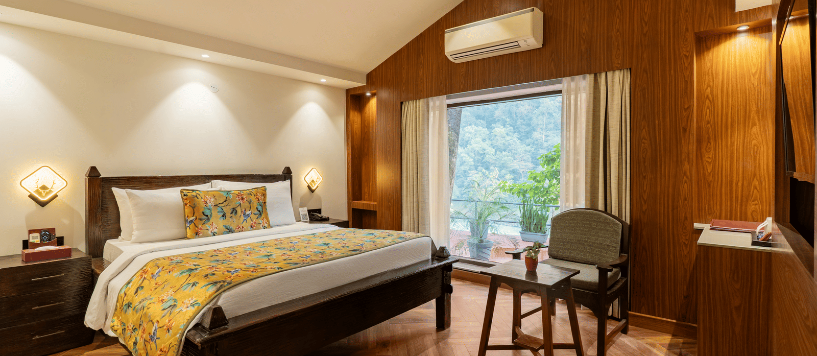 A spacious bedroom featuring a wooden bed, 2 bedside cabinets, a wooden chair facing a small table, and attached balcony with potted plants overlooking the trees outside | Summit by the Ganges Beach Resort & Spa, Rishikesh