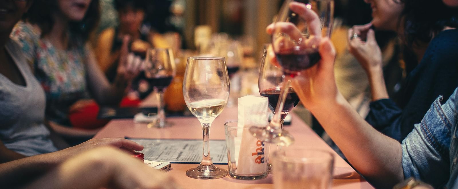 A long row of people seated at a table, having drinks, visibly wine, and bar snacks.