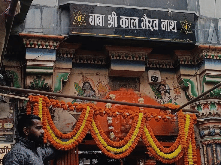 A street-level view of a small temple entrance decorated with orange flower garlands and a sign | HHI Varanasi