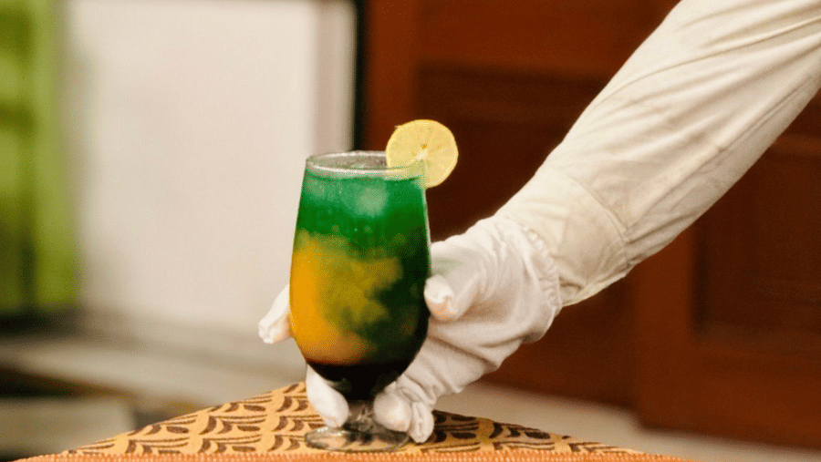 A person wearing a white glove holds a glass containing a layered green and yellow drink at Amantra Shilpi Resort, Udaipur.