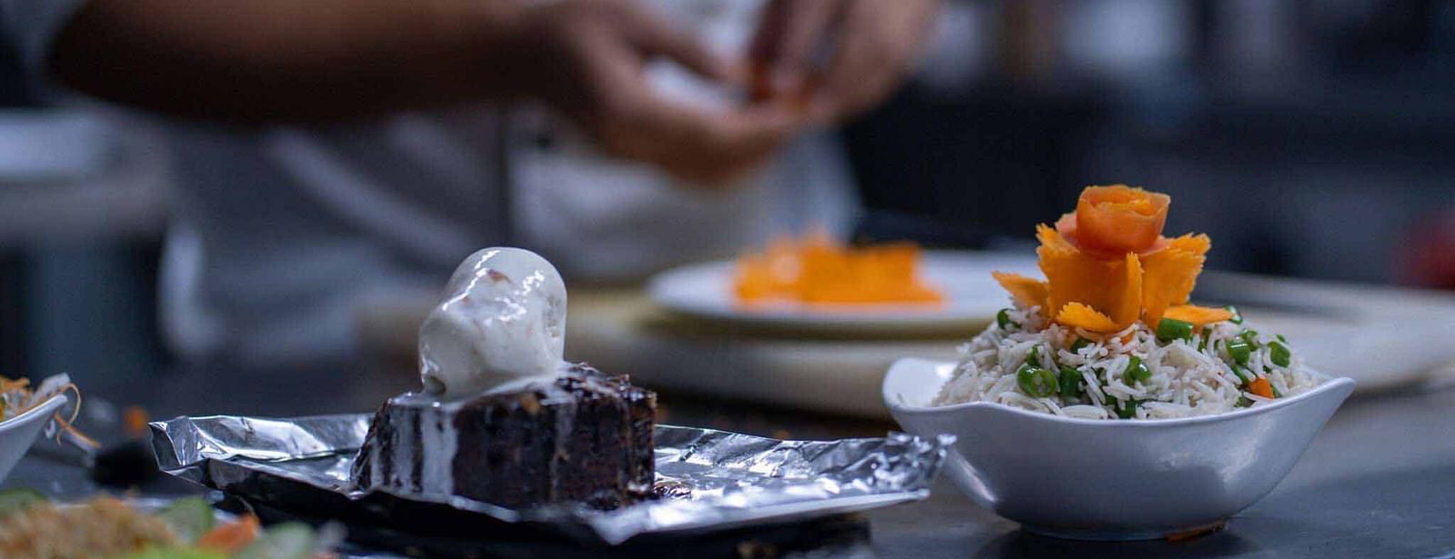 A blurred close-up shot of a chef's hands preparing dishes, with other plated items nearby - Mansha Hotel Regency