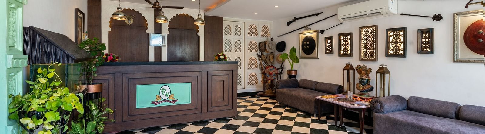 Reception area at Panna Vilas Palace features a wooden reception desk, walls adorned with wall-mounted frames, and several potted plants | Panna Vilas Udaipur