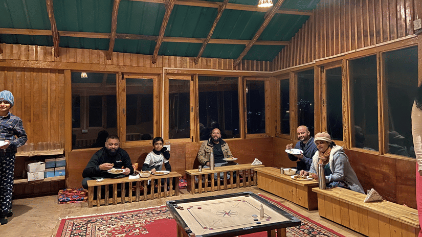 A cosy dining space at Amara Upepo - The Sky Village featuring several people having a meal dressed in winter-wear, along with a carrom board in the middle of the room.