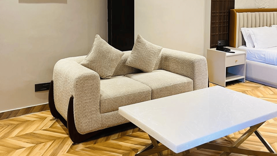 A hotel room corner featuring a beige sofa with cushions, a marble-top coffee table, and patterned wooden flooring. The wall behind the sofa displays a modern abstract artwork, illuminated by recessed ceiling lights - Silver Sand Beach Resort, Havelock