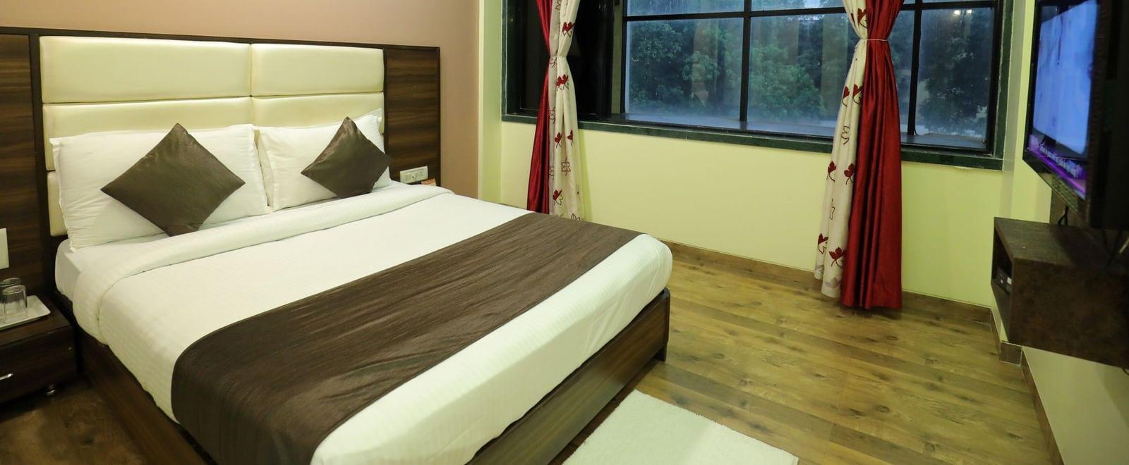 An Executive Room at Krushnai Resort with a white bed, brown accents, and a window with red curtains.