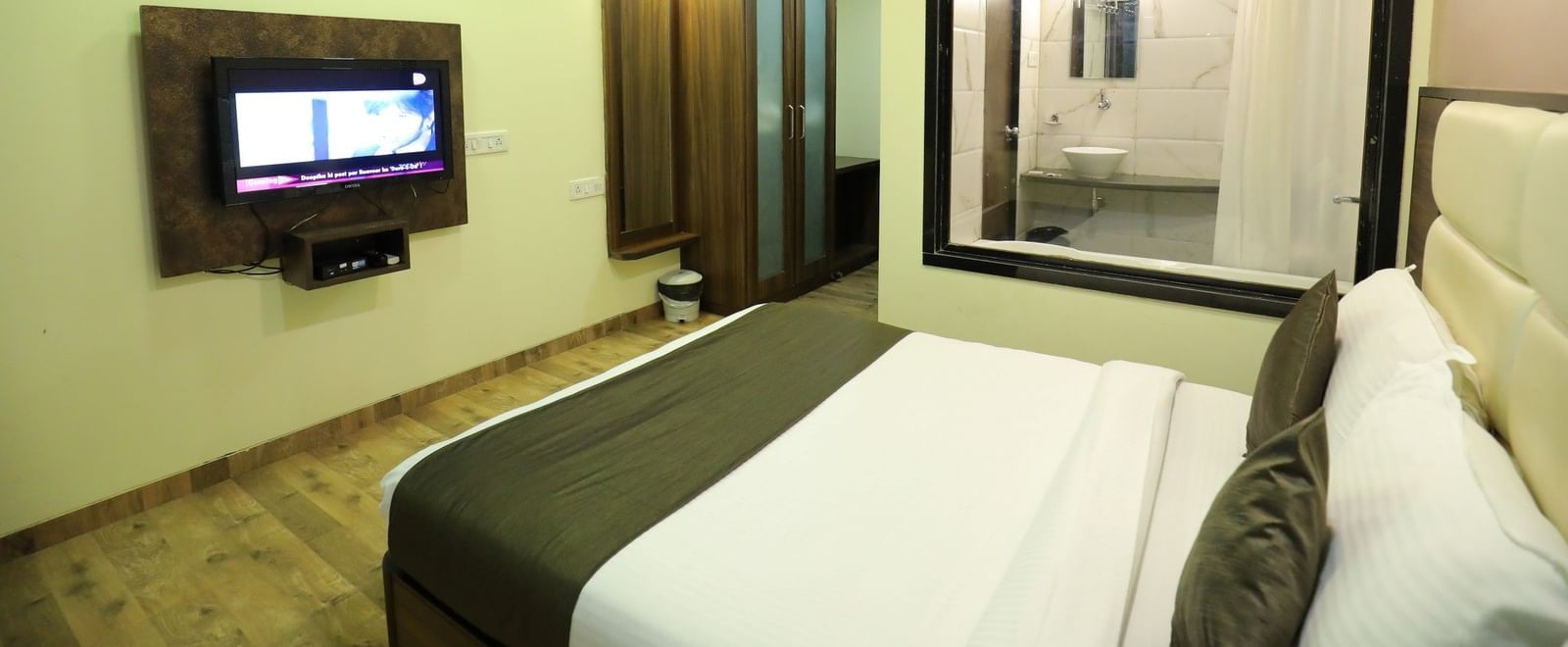 A bedroom at Krushnai Resort with a wall-mounted TV, a bed, and a glass-enclosed bathroom.