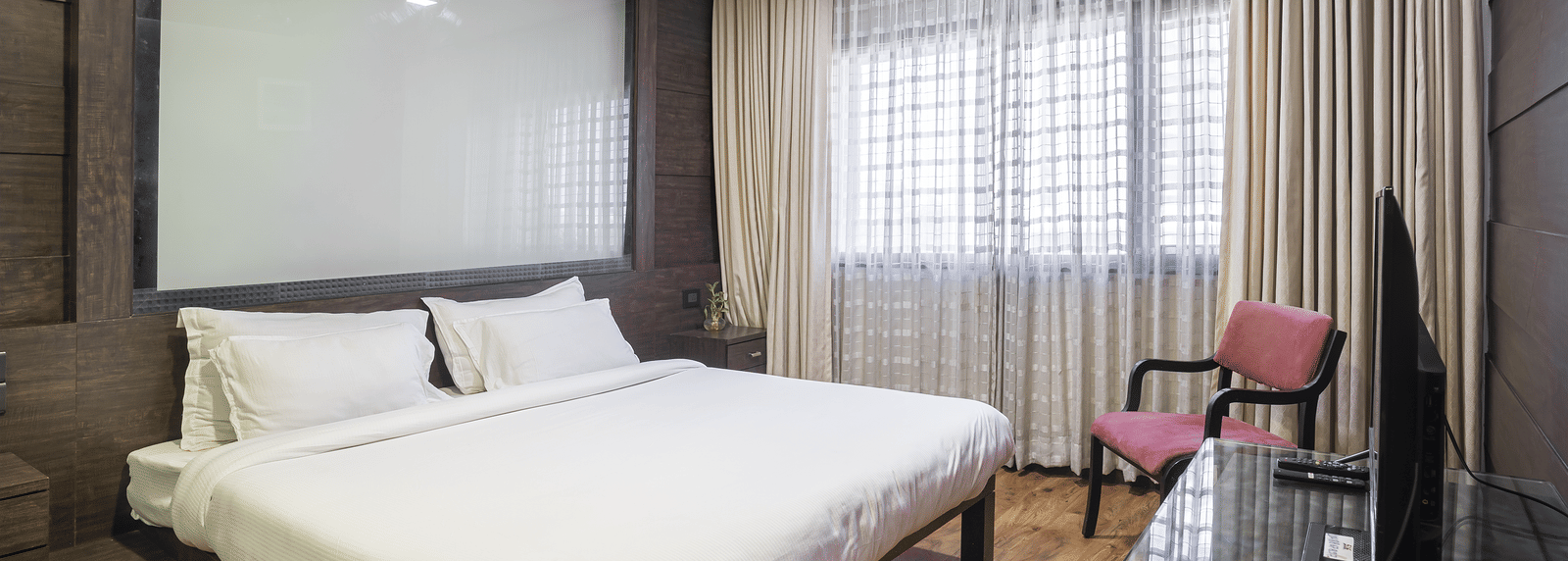 A spacious room with a king size bed, beige curtains, dark wall panelling, a pink armchair, and a T.V. at Infiniti Hotel, Indore.
