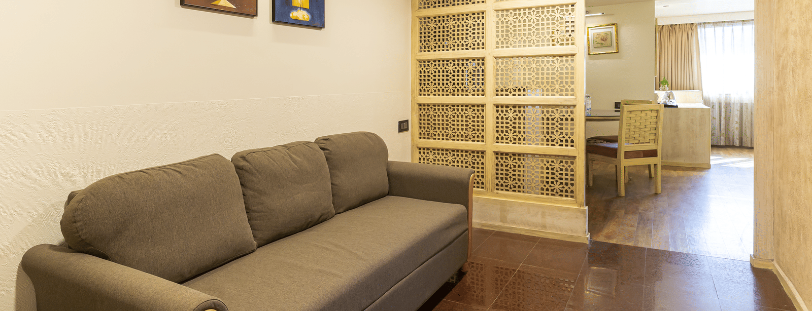 Room with a grey sofa, wooden floor, paintings on the wall and a decorative panel in the background at Infiniti Hotel, Indore.