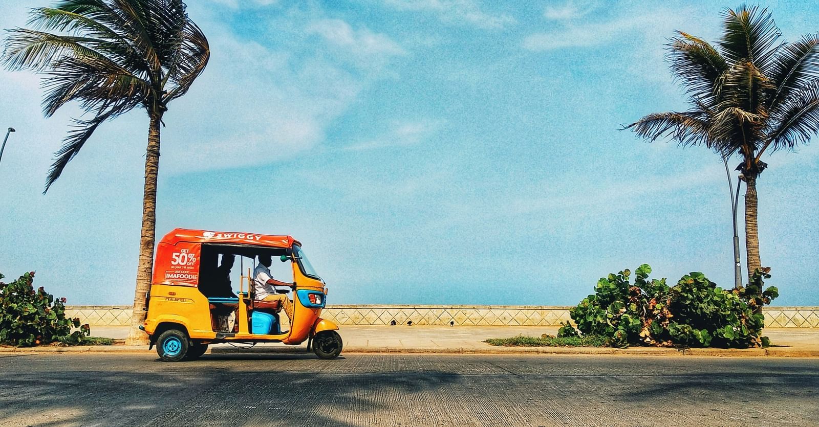 A yellow and orange auto-rickshaw is parked on a paved road next to a sandy beach.