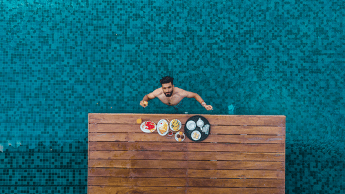 Aerial view at Symphony Oceanus Bay Beach Resort & Spa of a man swimming up to a pool ledge set with a breakfast tray of fruit, waffles, bananas, and tea.