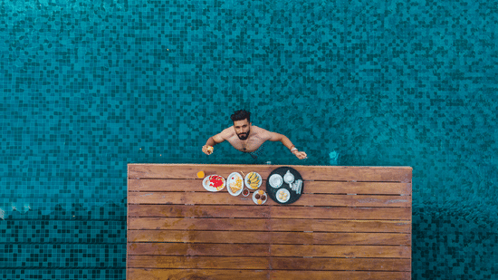 Aerial view at Symphony Oceanus Bay Beach Resort & Spa of a man swimming up to a pool ledge set with a breakfast tray of fruit, waffles, bananas, and tea.
