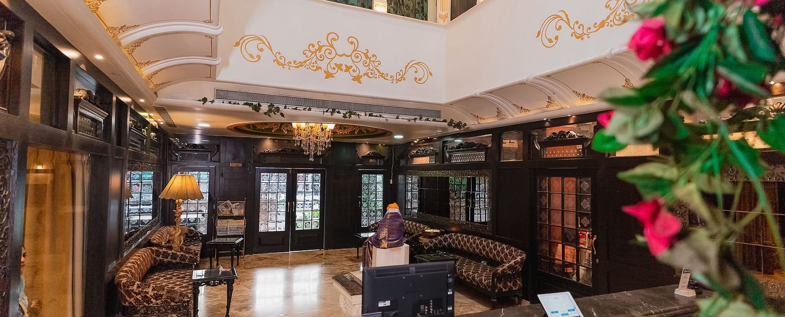 Hotel Ashok Nagpur featuring a wooden check-in counter with a computer and other equipment, a blurred figure in the background, and a floral arrangement on the side.