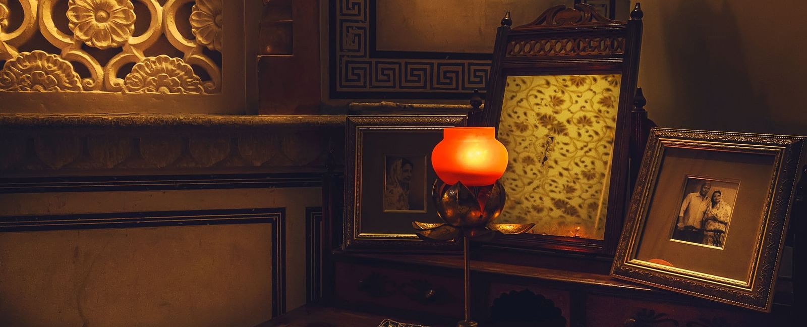 A dimly lit room at Umaid Vilas Jaipur featuring a decorative wall cutout, a lamp with an orange shade, and framed artwork.