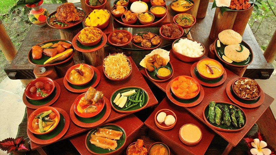 A vibrant festive food spread at Vedic Village Spa Resort featuring multiple traditional dishes served in clay and wooden bowls arranged on a decorated table with tropical foliage.