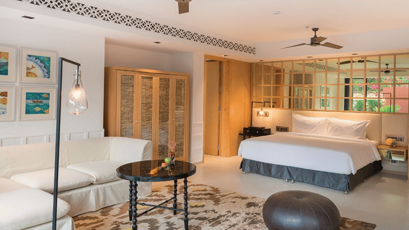 The private pool villa in Goa with a pouffe and a coffee table surrounded by a white couch in front of a plush bed at Azaya Beach Resort Goa