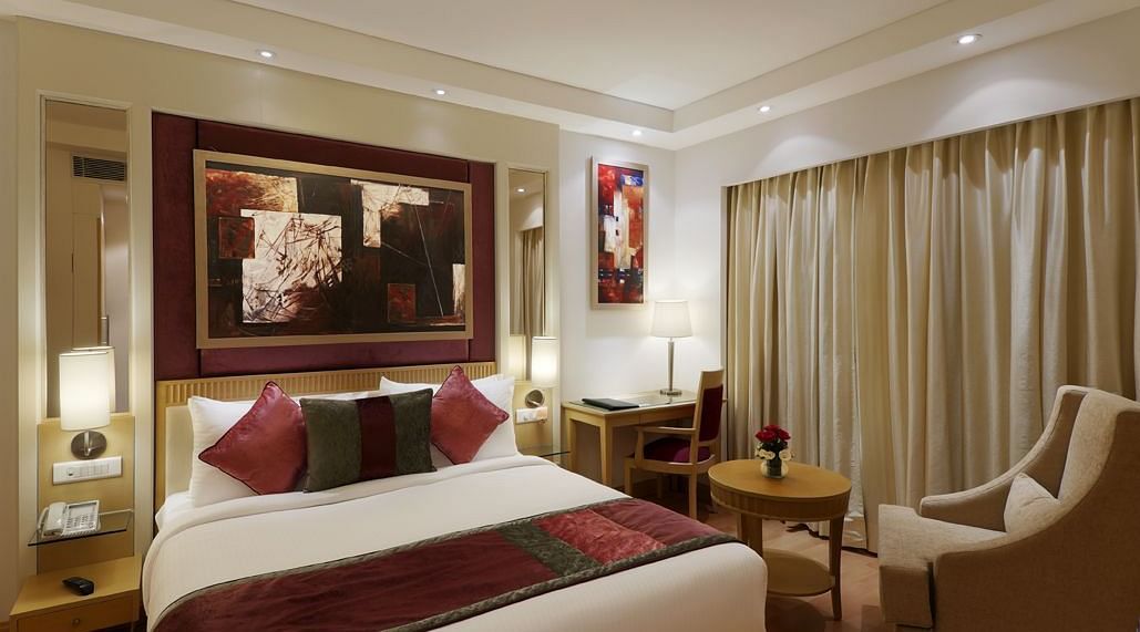 A beautifully designed bedroom featuring a double bed, a sofa chair with a small coffee table, and wall decor such as paintings – Inde Hotel Vista Woods, Gurgaon.