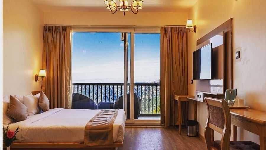 A sunlit bedroom with a white and gold bed, a wooden platform, and a large window framing an outdoor landscape - Clarks Exotica Resort, Near Mukhteshwar.