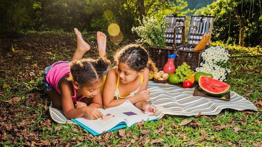 Two girls lying under the sun on a mat with picnic spread on the side and smiling while reading a book