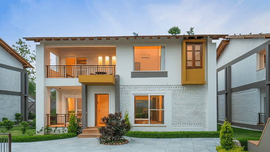 A two-story villa with a balcony, surrounded by lush greenery and a clear blue sky.