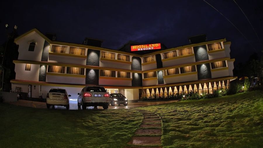 Facade of Highland Resort at night, featuring park cars in front, a large illuminated signboard of the resort, and a well-lit multi-storey building with lawn in front.