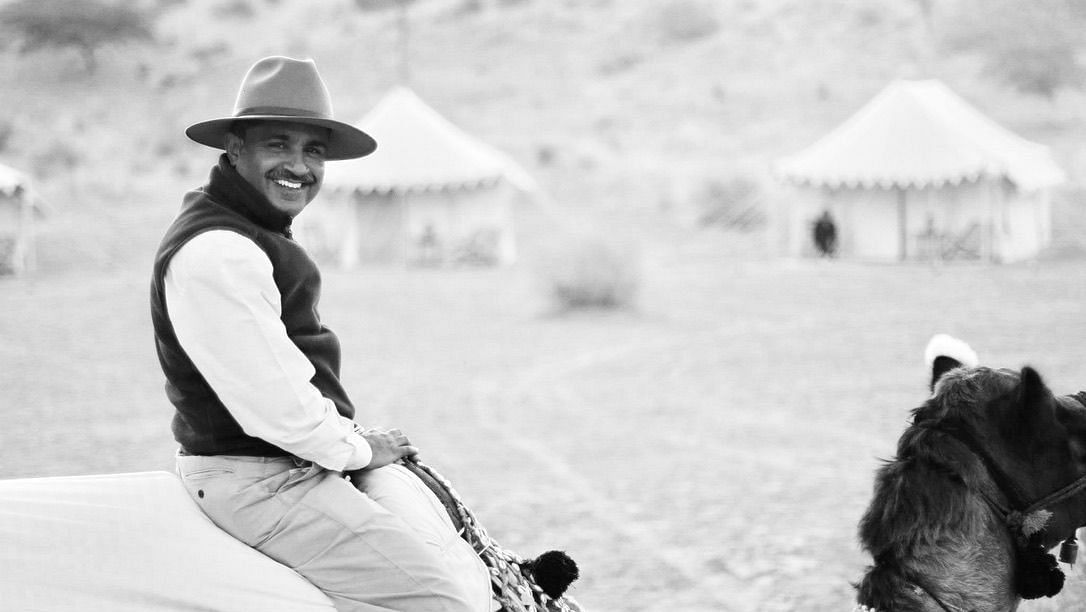 A black and white image of a man riding a camel with two tents in the background - MANVAR Resort & Desert Camp