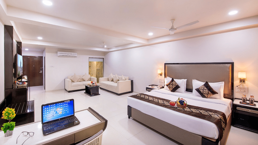 A comfortable hotel suite offers a king size bed, a separate seating area, a functional desk with a laptop, and ample natural light.