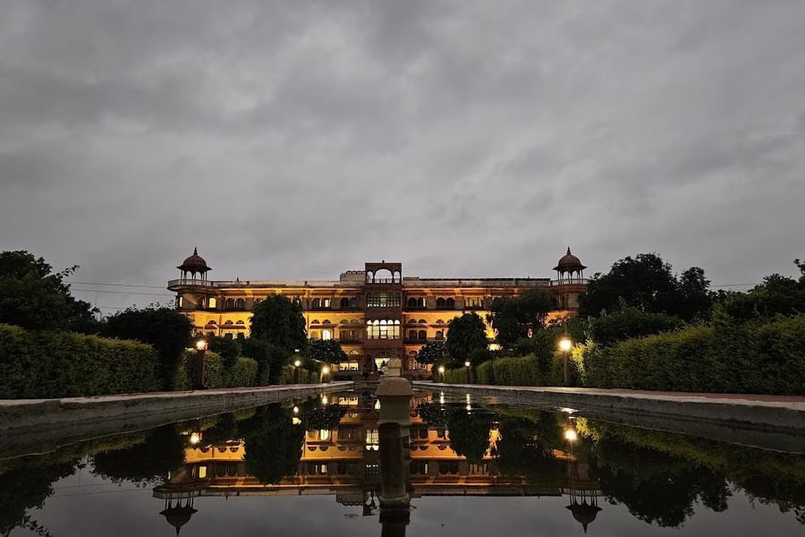 Well lit facade of Umaid Palace Dausa featuring stretched and multi storey building with lights on. The property is covered by trees and there is also a  huge fountain in the front yard.
