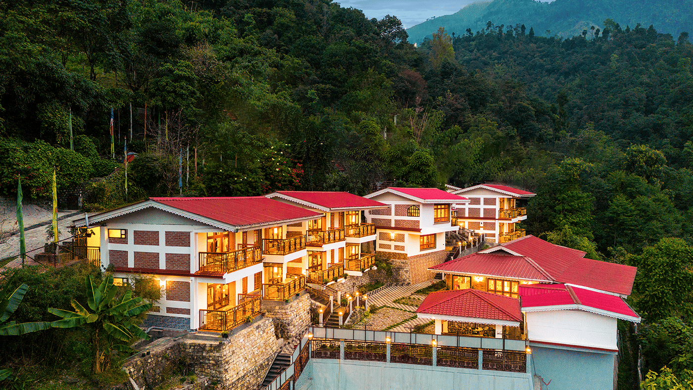 A hillside voyage songfum retreat with multiple buildings featuring red roofs and glowing lights, surrounded by dense green forest and mountains in the background.