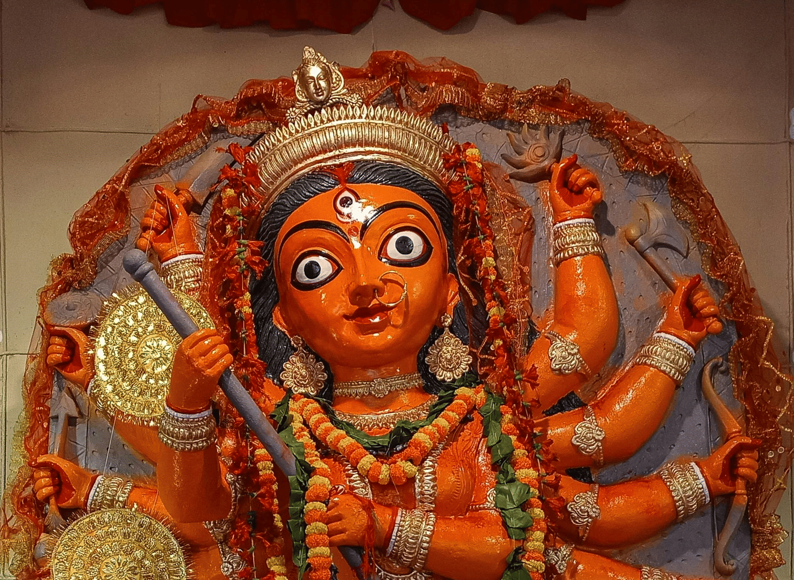 Statue of a Goddess Durga with multiple arms, dressed in orange with golden coins in the background.