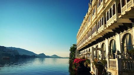 A multi-story building with arched windows and a balcony faces a calm body of water with distant hills.