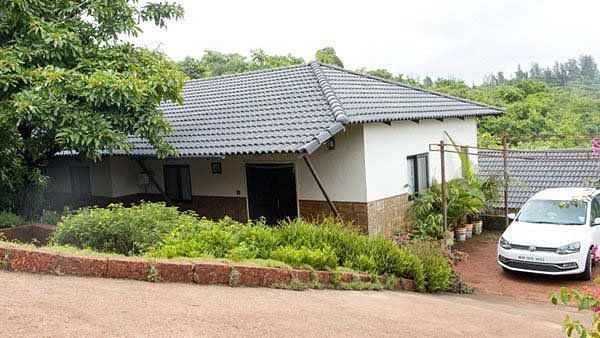 A single-story house with a dark roof and white walls, nestled among trees and greenery, with a white car parked beside it.