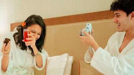 Two people sitting on a bed in robes, smiling and taking pictures with vintage-style instant cameras at Karma Chalets.