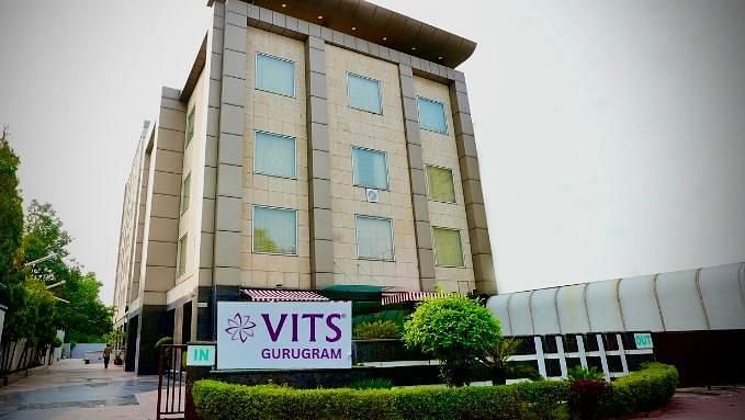 A low-angle exterior shot of a multi-storey building with a sign that reads VITS GURUGRAM, surrounded by greenery
