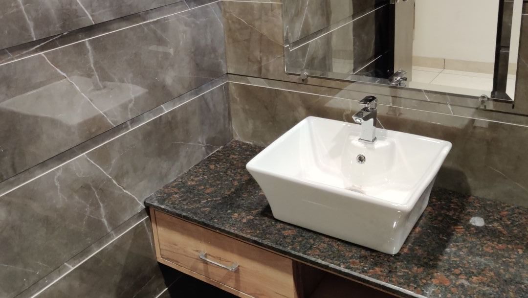 A close-up view of a bathroom at Hotel Exotic Amritsar with a textured grey tiled wall, a square vessel sink on a dark wood vanity, and a large rectangular mirror.