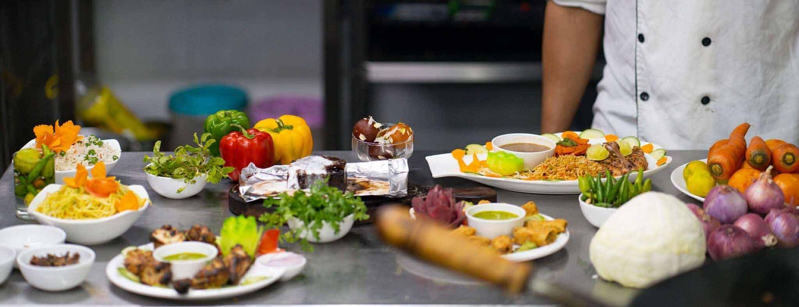A chef in a white uniform standing behind a counter laden with various ingredients, fresh produce, and prepared dishes at Mansha Hotel Regency.