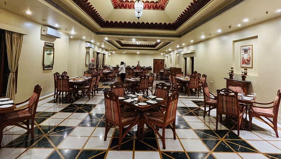 A spacious dining space with ornate ceilings, chandeliers, and a checkered black and white floor. Round tables with chairs are arranged throughout the hall, and there are staff members setting up for an event.TUJ - Jharokha Restaurant