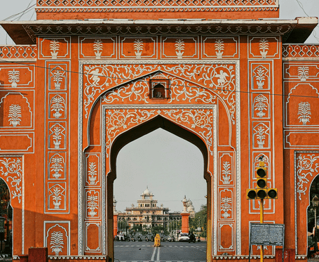 A grand, intricately decorated orange archway frames a view of a building in the distance under a pale sky.