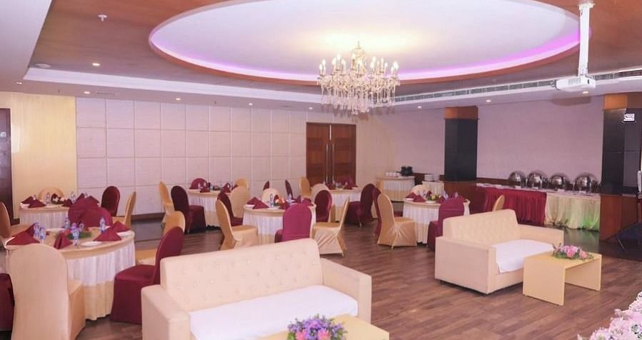 A banquet hall in Dhanalakshmi Srinivasan Hotels, Perambalur, with round tables, chairs, sofas, and a floral arrangement in the foreground. The hall has wooden flooring and a chandelier.