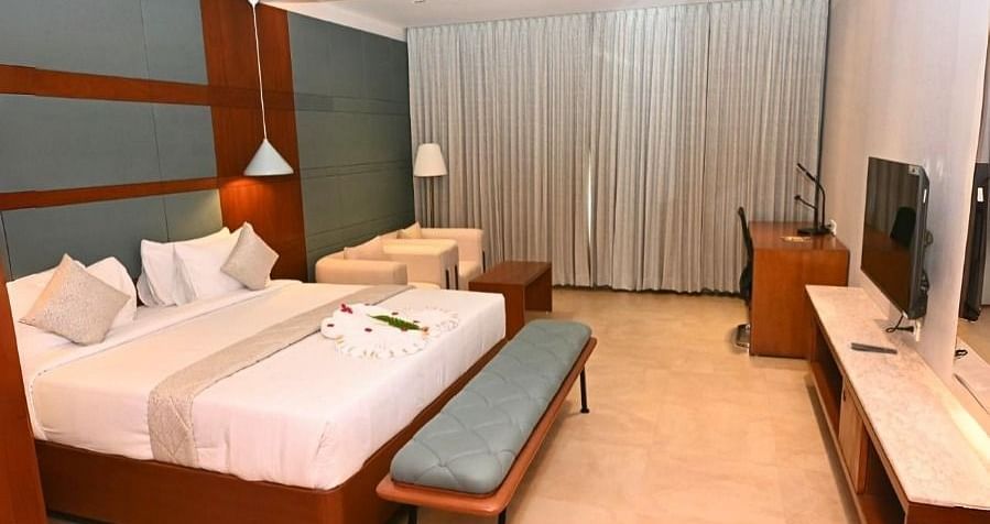 A hotel room at Dhanalakshmi Srinivasan Hotels, Perambalur, featuring a double bed, two benches, a TV on a console, a desk with a chair, two armchairs, and closed curtains.