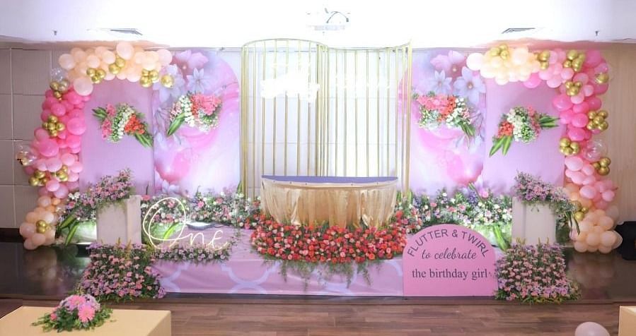 A decorated hall in Dhanalakshmi Srinivasan Hotels, Perambalur, set up for a celebration with a stage adorned with pink and gold balloons, flowers, and a banner. There are sofas and a coffee table in the foreground.