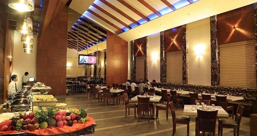 The interior of the Aroma restaurant in Dhanalakshmi Srinivasan Hotels, Perambalur, showing a buffet spread, dining tables, and chairs. The restaurant features wooden ceiling beams and tiled flooring.