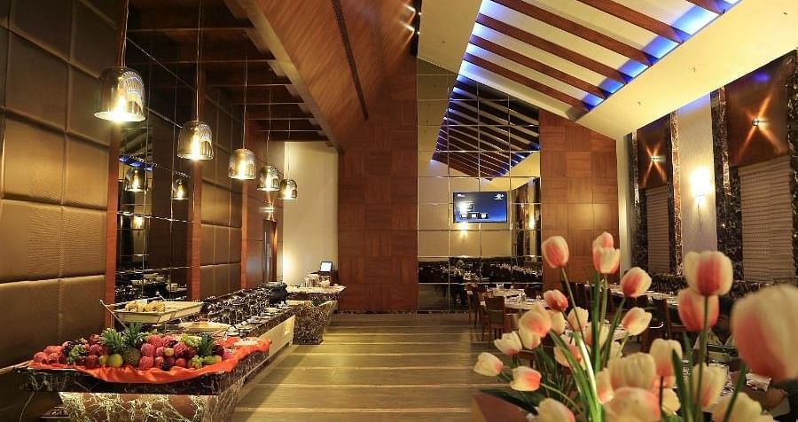A view of the Aroma restaurant in Dhanalakshmi Srinivasan Hotels, Perambalur, featuring a buffet counter on the left and dining tables in the background. The restaurant has wooden ceiling beams and a floral arrangement in the foreground.