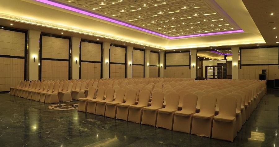 A large empty hall in Dhanalakshmi Srinivasan Hotels, Perambalur, with rows of chairs facing a stage. The hall has dark marble flooring and a patterned ceiling with recessed lighting.